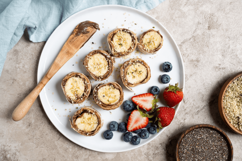 The image features banana sushi rolls on a white plate. There is a wooden spatula on the plate with a smear of sesame chocolate spread. There are also blueberries and whole and sliced strawberries on the plate. A blue scarf is to the left of the image.