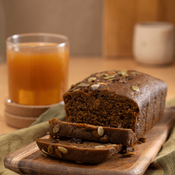Sliced Farm Boy Pumpkin Spice Loaf Cake on a wooden cutting board with glass of Farm Boy Sweet Apple Cider in background.