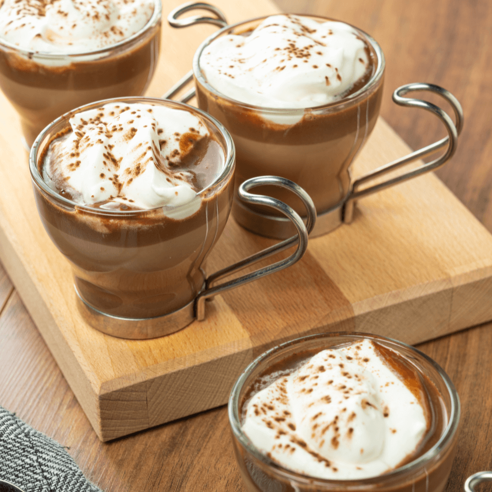 Four clear glass mugs of hot chocolate topped with whipped cream and cocoa powder on wooden cutting board.