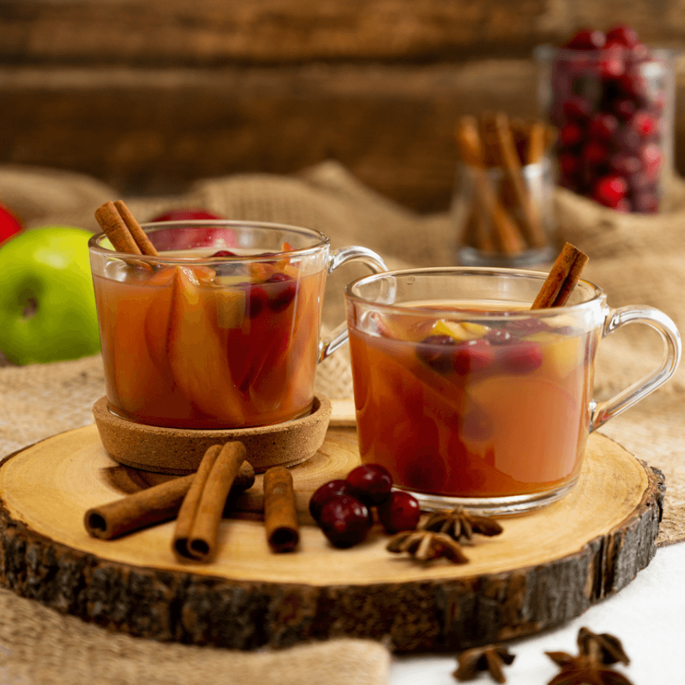 Two clear glass mugs of mulled apple cider with cranberries, apples, and cinnamon sticks on a live-edge wooden serving board.